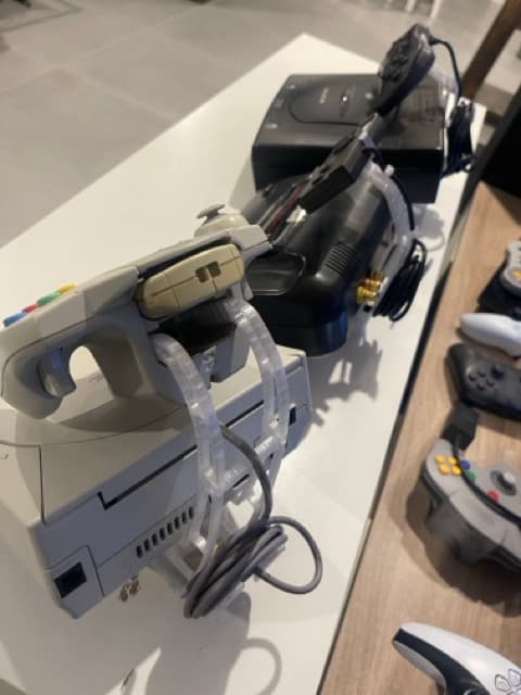 Printed clips and stands holding a light gun and its cable on a retro console display shelf, with N64, GameCube and Dreamcast controllers alongside