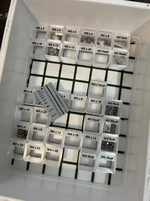 A tray of small labelled bins holding M2, M3 and M4 screws, nuts, washers and heat-set inserts, with a printed screw gauge resting on top